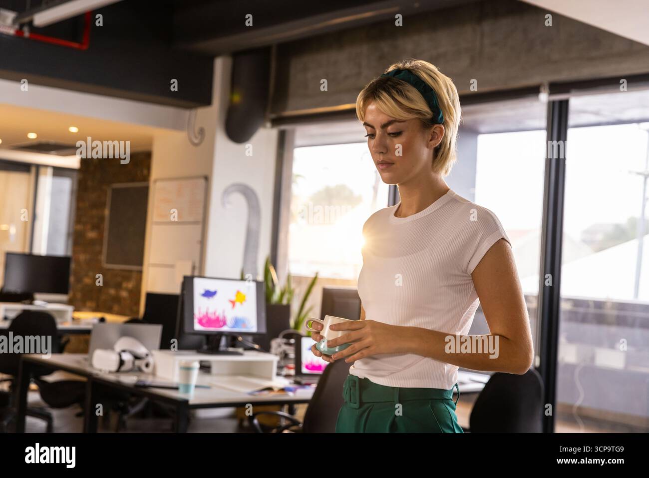 Donna in piedi nello spazio di lavoro dell'ufficio con tazza in ceramica che esamina i monitor del computer e copia lo spazio Foto Stock