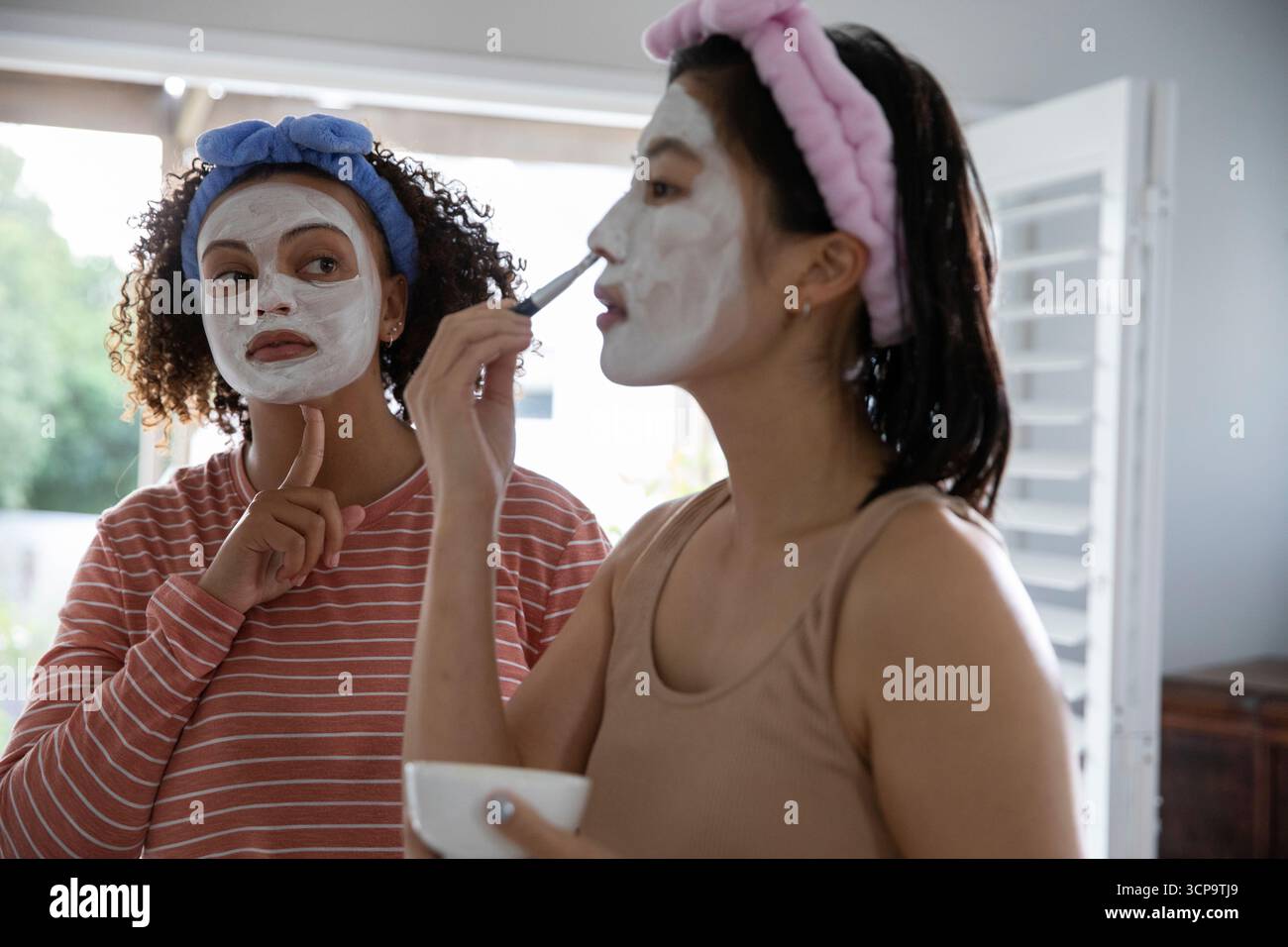 Diverse amiche che indossano morbide fasce alla vanità applicando maschere facciali con spazzola e ciotola Foto Stock