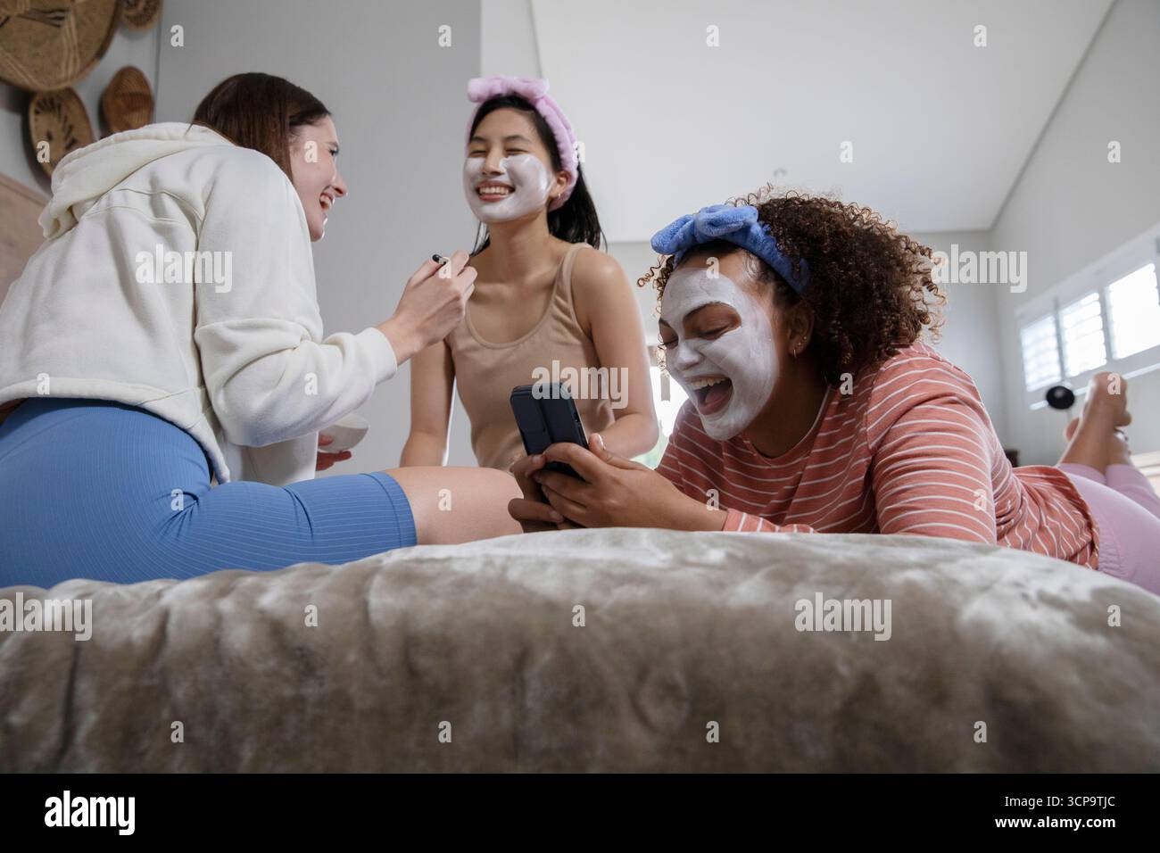 Diverse amiche che si rilassano in camera da letto applicando maschere facciali con pennello e tenendo in mano lo smartphone Foto Stock