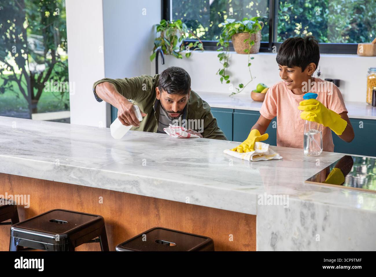 Padre dell'asia meridionale con figlio che pulisce il piano di appoggio in marmo dell'isola della cucina usando bottiglie spray Foto Stock