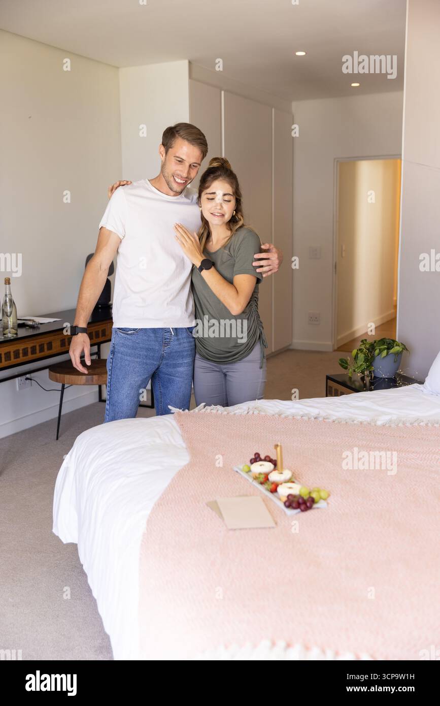 Coppia in piedi accanto al letto nella camera da letto di casa, tenendosi l'un l'altro ed esaminando il vassoio della colazione Foto Stock