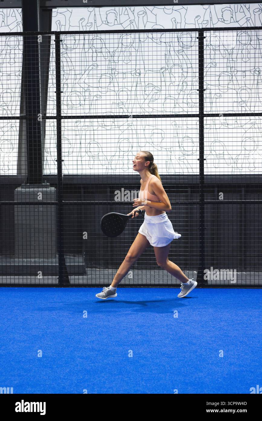 Atleta che indossa un abbigliamento sportivo rosa running forward che colpisce con la racchetta sul campo blu del padel Foto Stock
