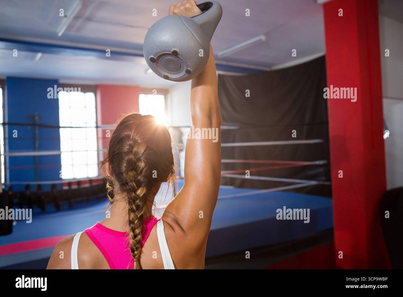 Donna che indossa la parte superiore rosa e i leggings che sollevano il kettlebell grigio nell'anello di boxe con grandi finestre Foto Stock