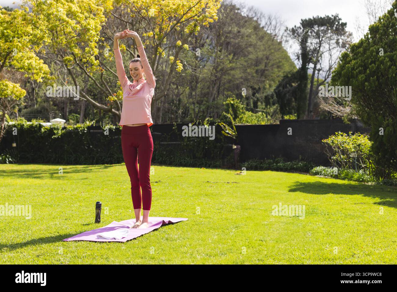 Donna che allunga le braccia indossando abiti da ginnastica su un tappetino yoga alla lavanda nel cortile accanto alla bottiglia d'acqua nera Foto Stock