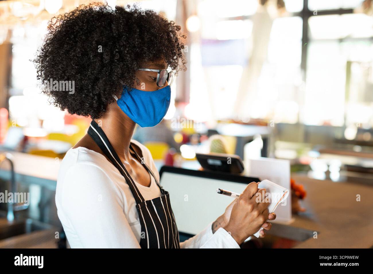 Donna afroamericana in grembiule, maschera, prendere l'ordine sul blocco note dietro il registro del caffè, spazio copia Foto Stock