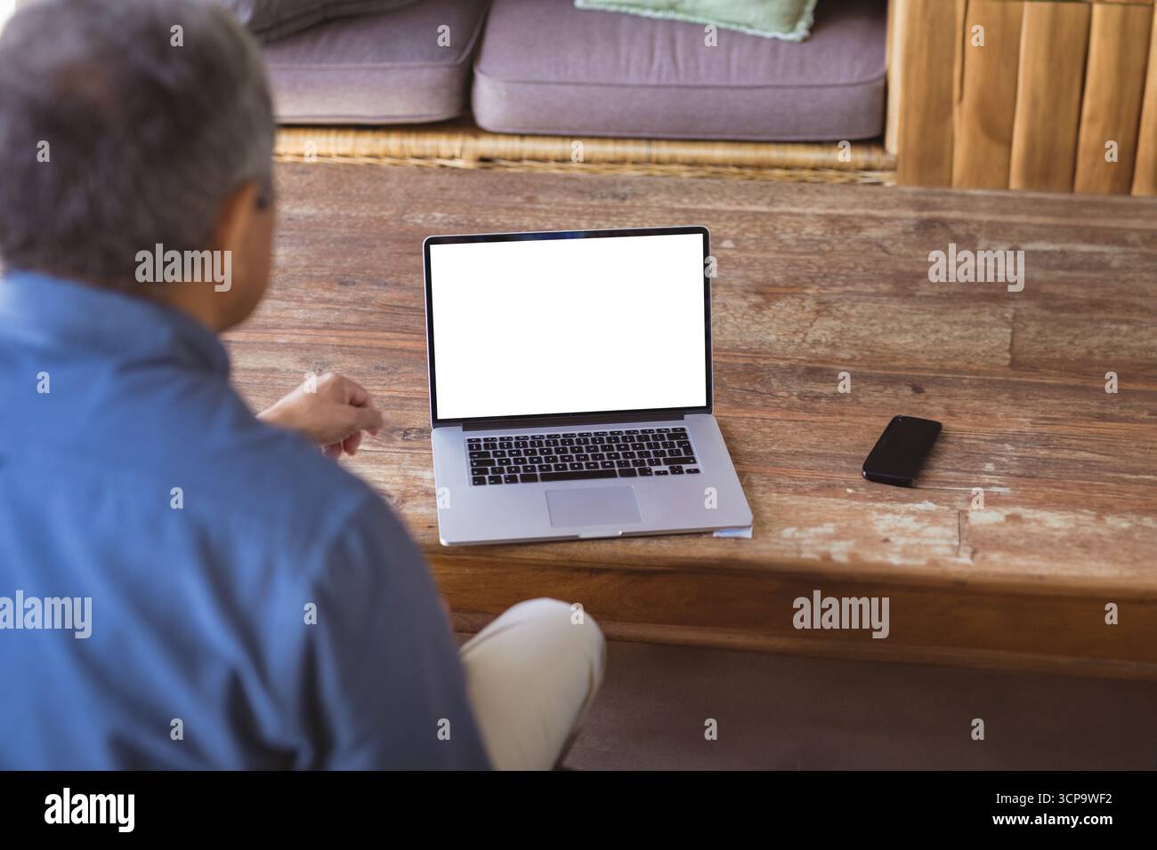 Uomo anziano seduto al tavolino da caffè di legno usando un computer portatile argentato con smartphone a casa Foto Stock