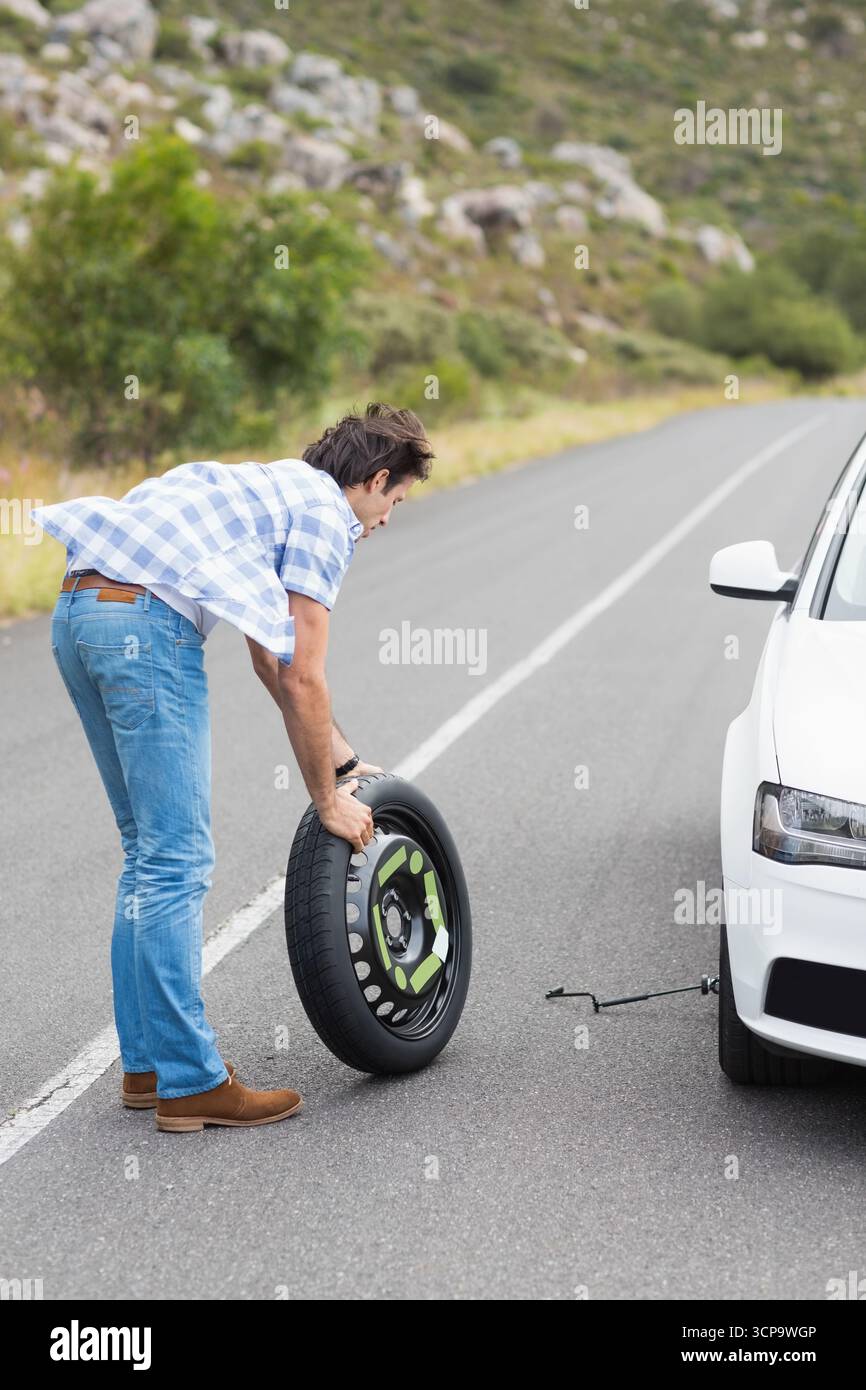 Uomo di mezza età che solleva la ruota di scorta accanto alla berlina su una strada di montagna utilizzando un martinetto e uno spazio per copiare Foto Stock