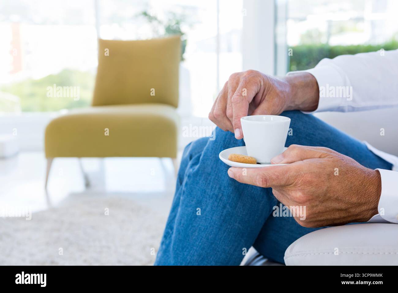 Le mani stanno tenendo una piccola tazza di caffè bianco con piattino e biscotti sul divano vicino alle grandi finestre Foto Stock