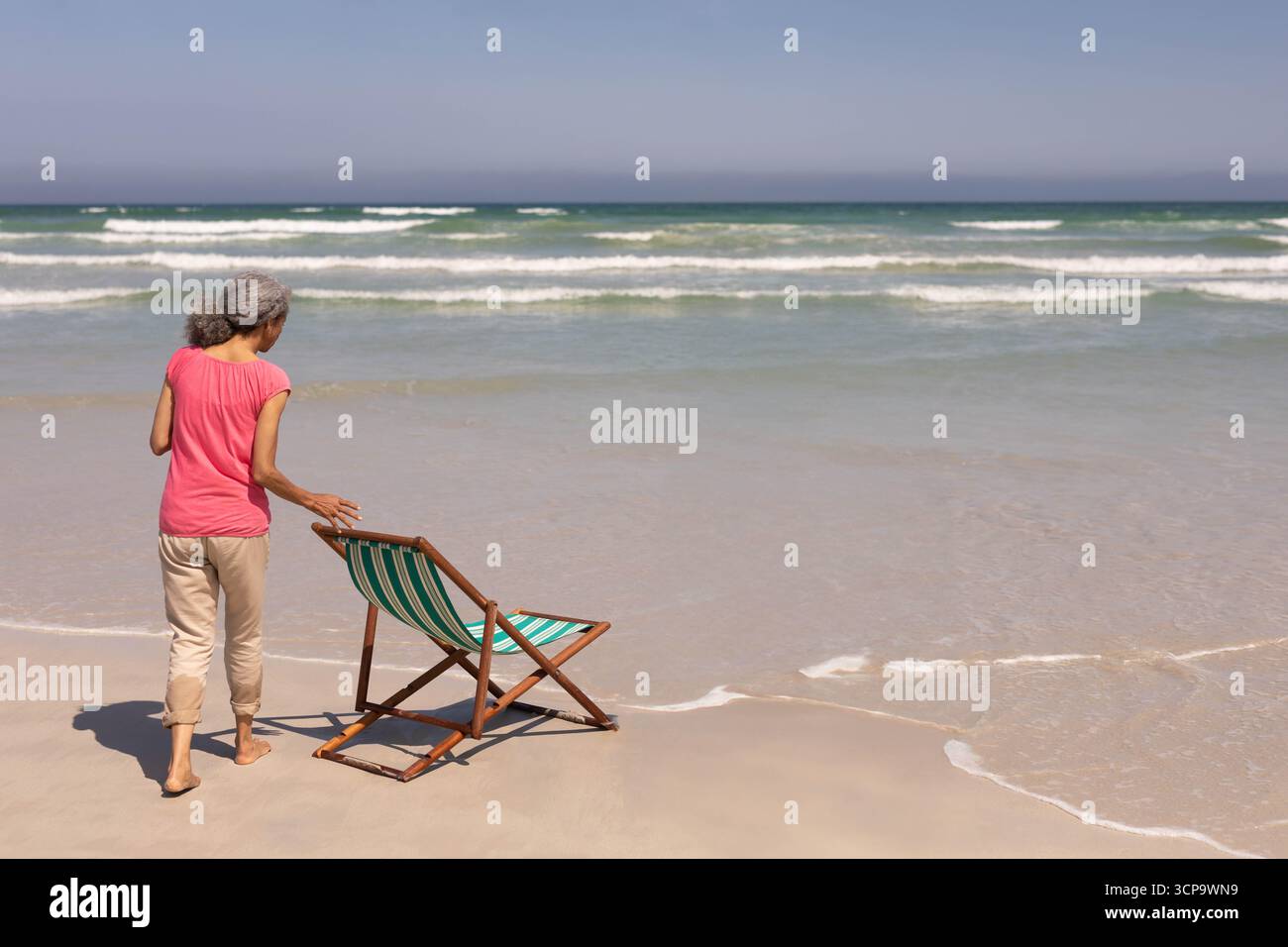Donna afroamericana senior che posiziona una sedia da spiaggia a righe sulla sabbia bagnata sul bordo dell'oceano, copia spazio Foto Stock