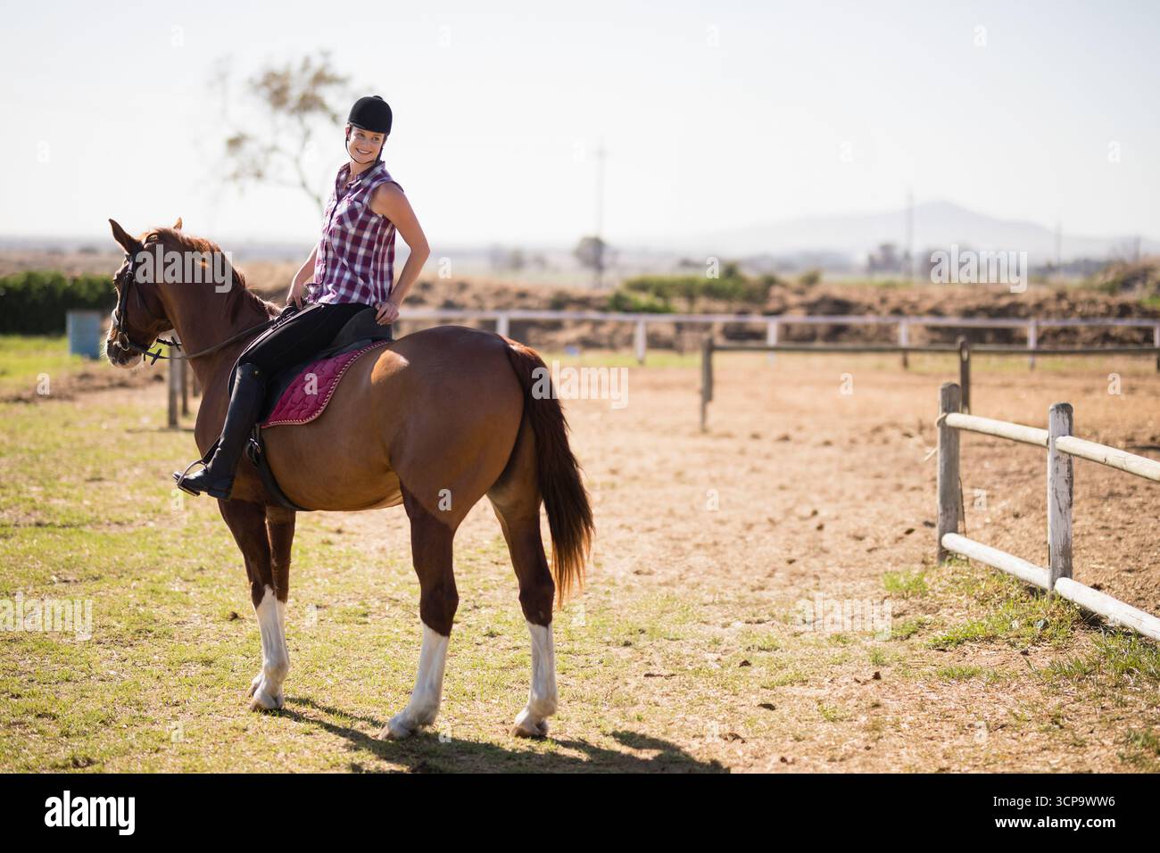 Pilota donna seduta a cavallo di castagno in un paddock di terra che indossa un casco con sottosella Bordeaux Foto Stock