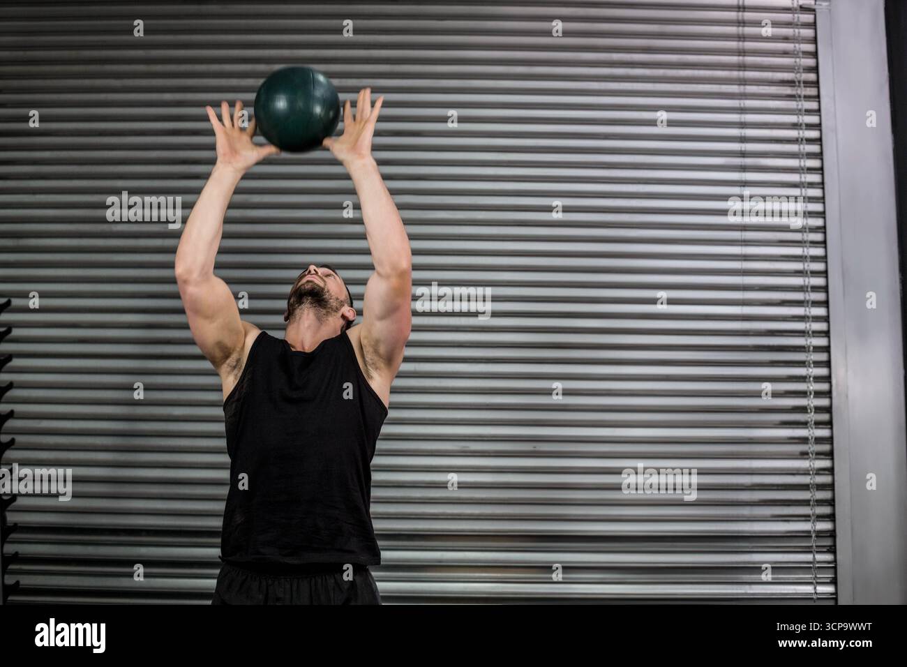 Atleta maschile che indossa una canotta nera che solleva una palla medica verde sopra la palestra accanto alla porta dell'otturatore Foto Stock