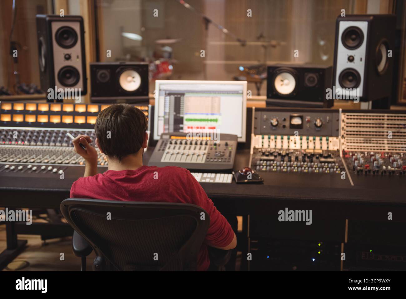 Ingegnere audio maschio che mixa tracce su console nella sala di controllo dello studio con kit batteria in background Foto Stock