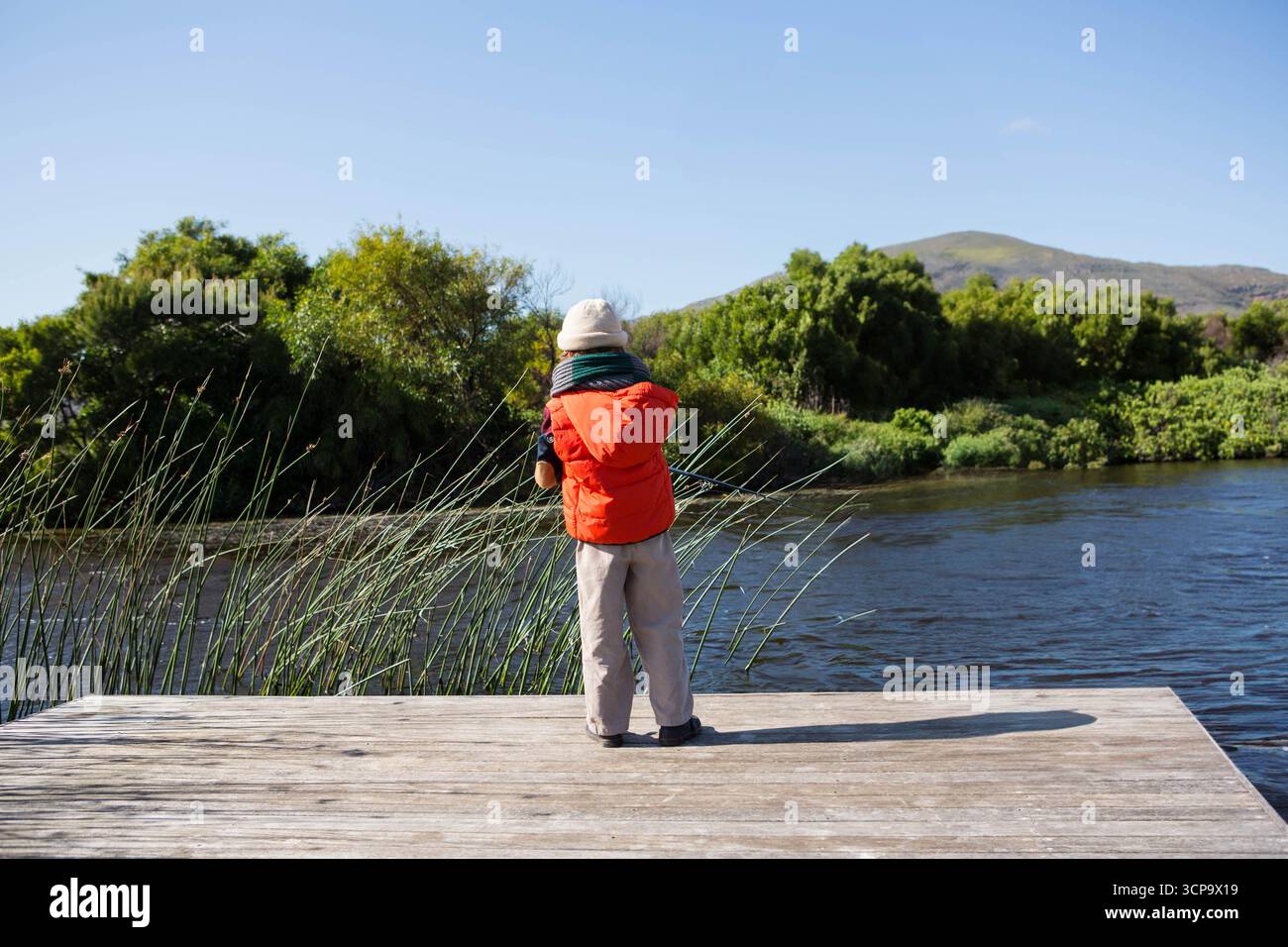 Ragazzo che indossa un gilet arancione e un cappello da secchio in piedi su un molo di legno che guarda il lago calmo Foto Stock