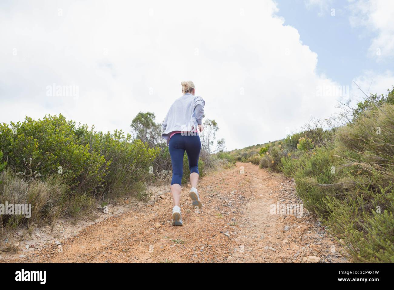 Donna adulta che corre in salita su un sentiero rurale con giacca grigia, leggings navy, scarpe da running Foto Stock