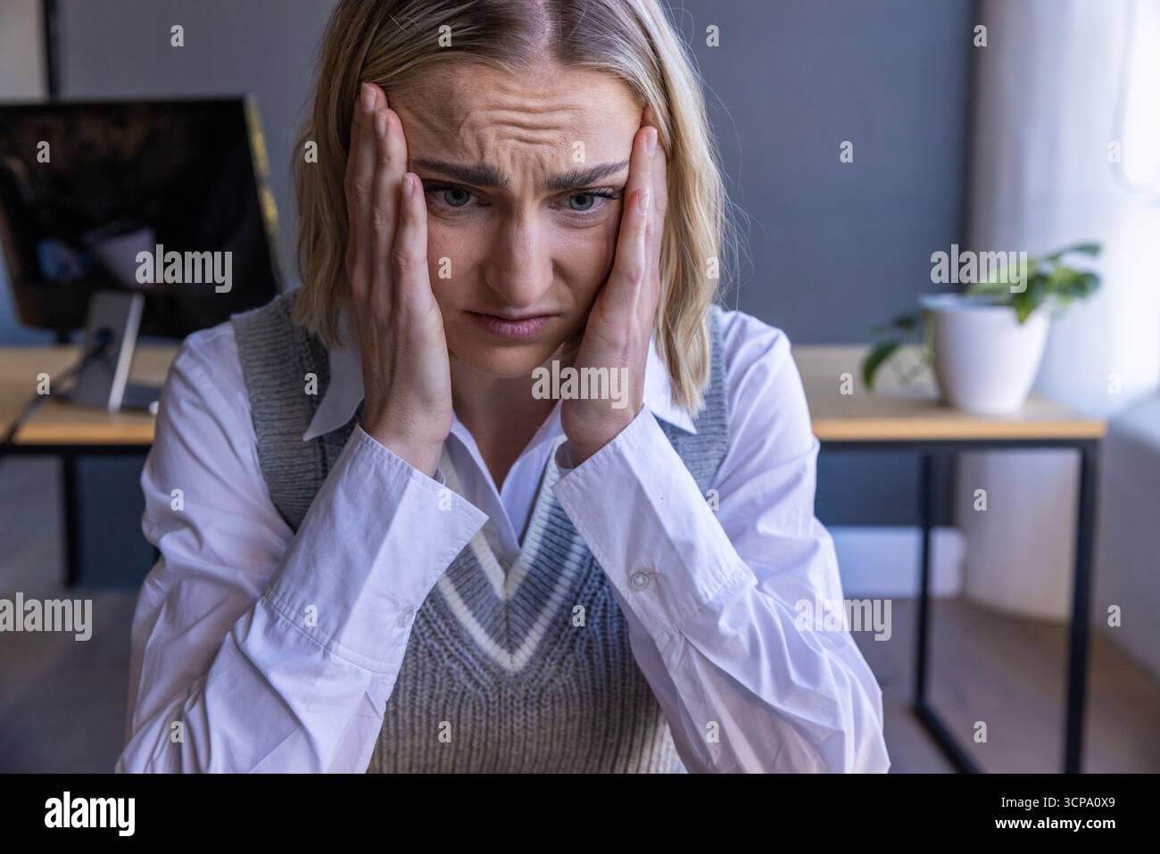 Donna che indossa un gilet grigio su una camicia bianca che tiene le aste alla scrivania dell'ufficio accanto al monitor Foto Stock