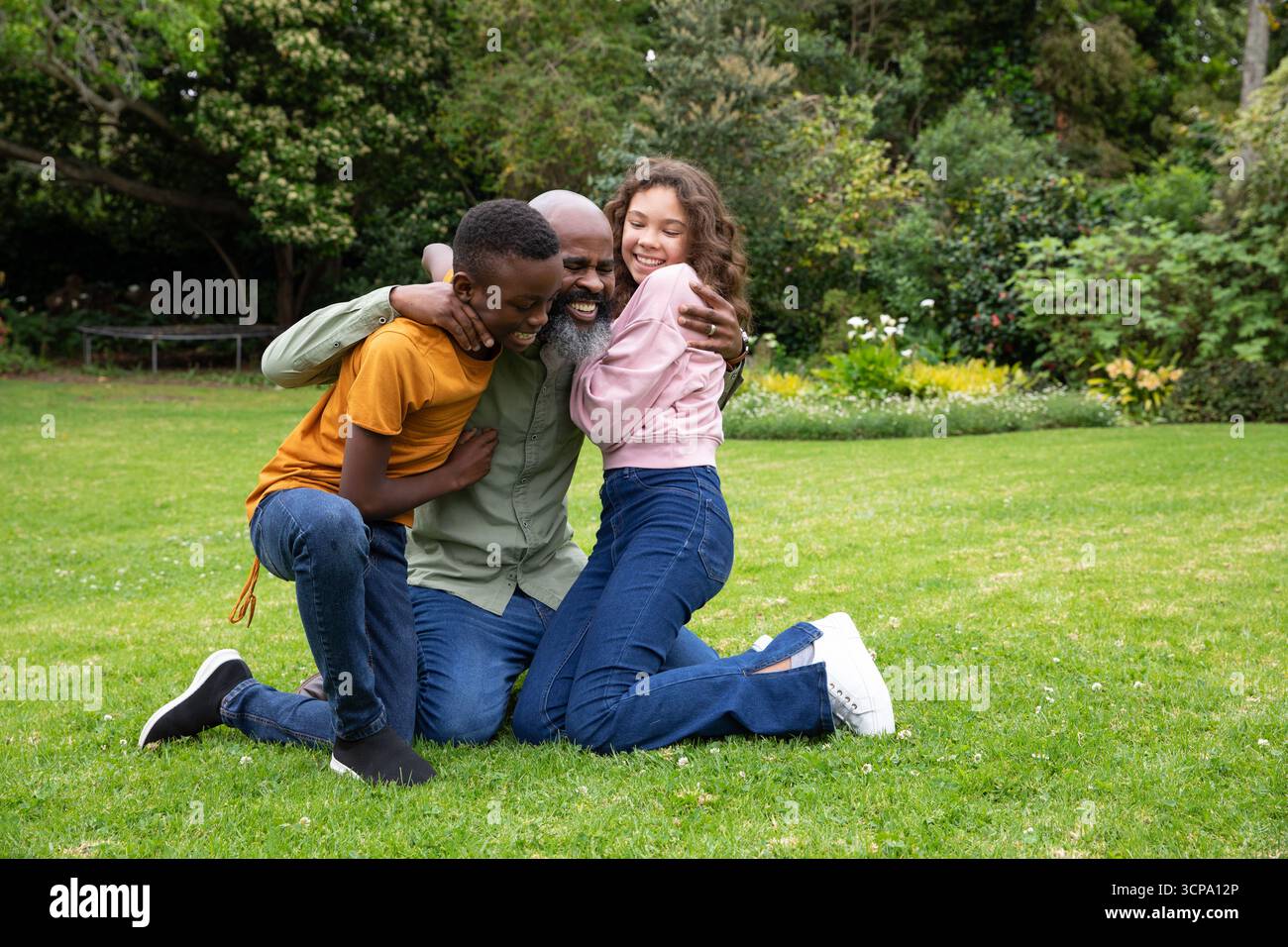 Famiglia afroamericana che abbraccia i bambini vicino mentre si inginocchia sull'erba vicino a aiuole di fiori Foto Stock