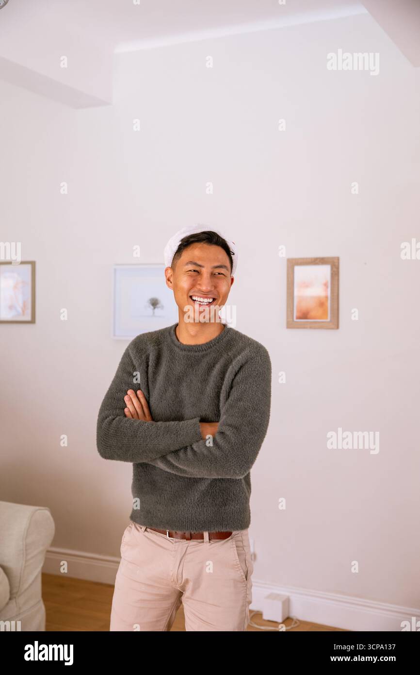 Uomo asiatico in piedi con le braccia incrociate nel soggiorno vicino al divano, arte incorniciata, adattatore elettronico Foto Stock
