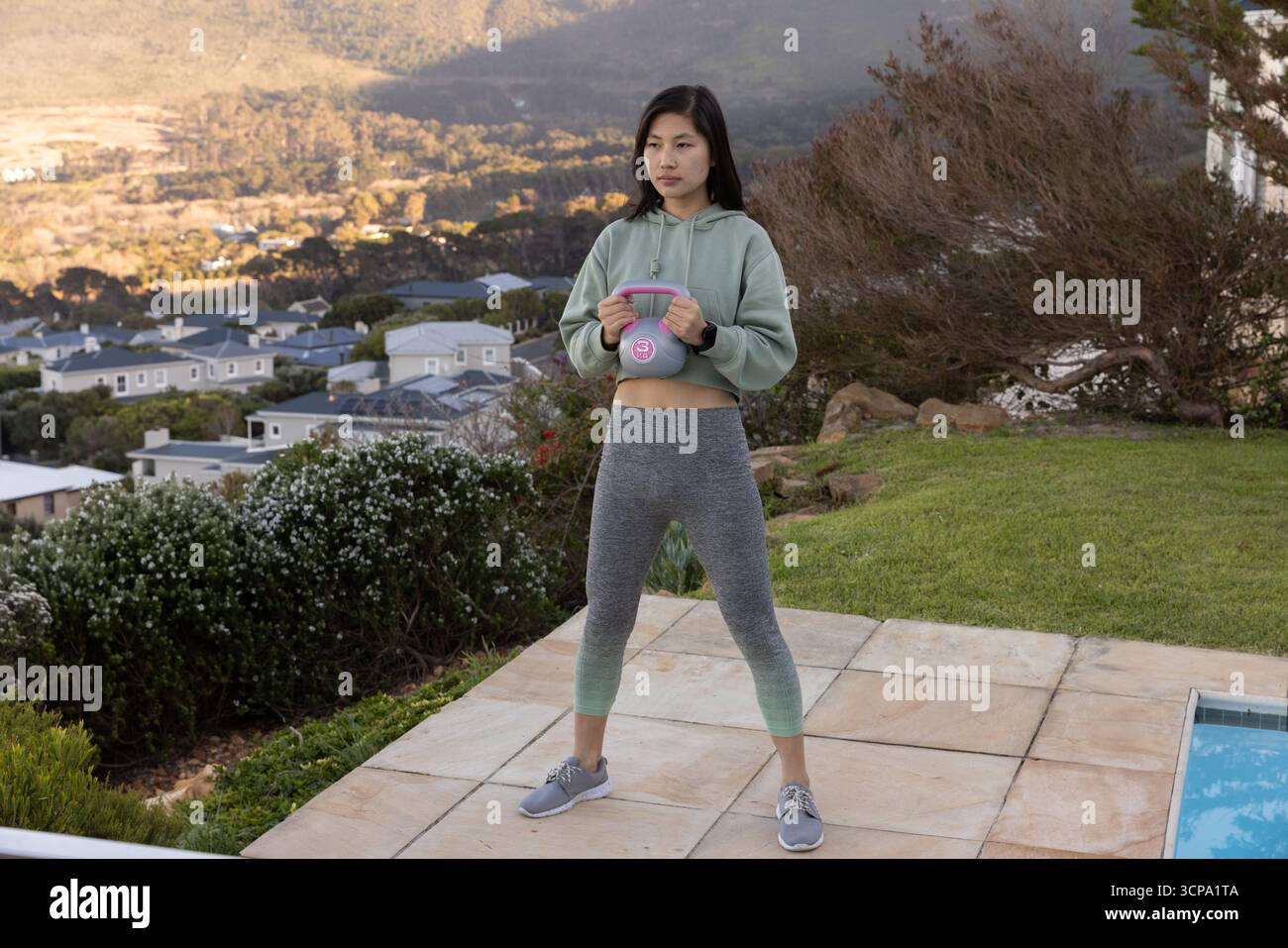 Donna asiatica che indossa l'attrezzatura da allenamento tenendo il campanello sul patio piastrellato accanto alla piscina Foto Stock