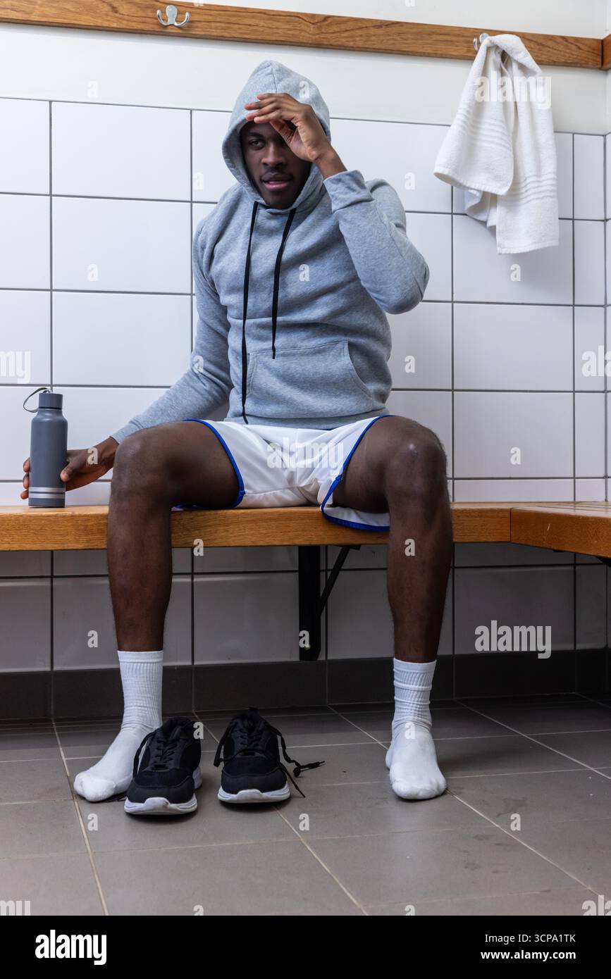 Uomo afroamericano seduto sulla panchina nello spogliatoio con cappuccio e in possesso di una bottiglia d'acqua Foto Stock
