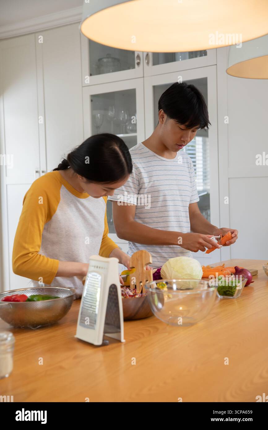 Coppia asiatica che taglia cavolo e peeling carote con pelapatate sull'isola in un'accogliente cucina Foto Stock