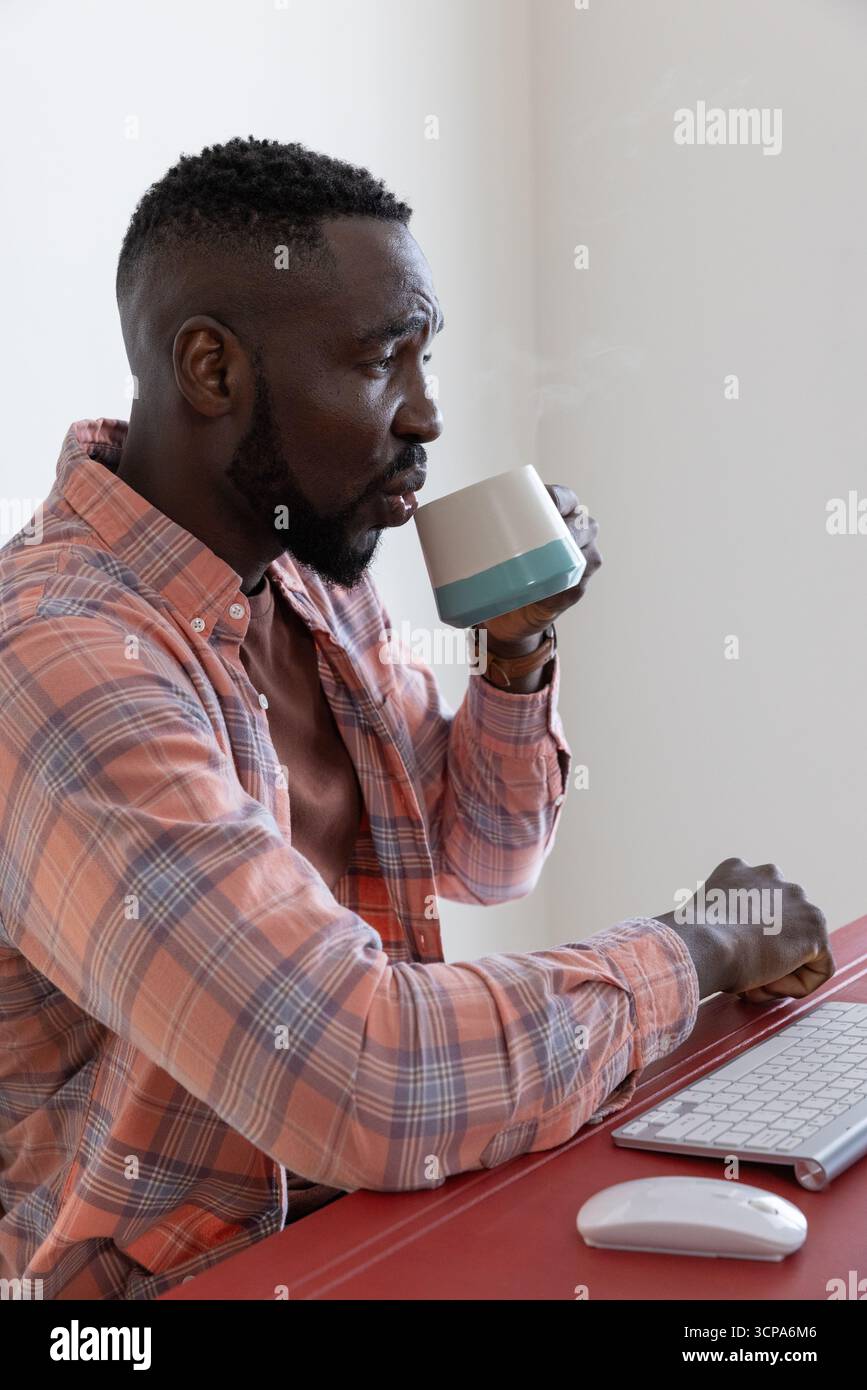 Uomo afroamericano che tiene in mano la tazza mentre lavora alla scrivania da casa con tastiera e mouse Foto Stock