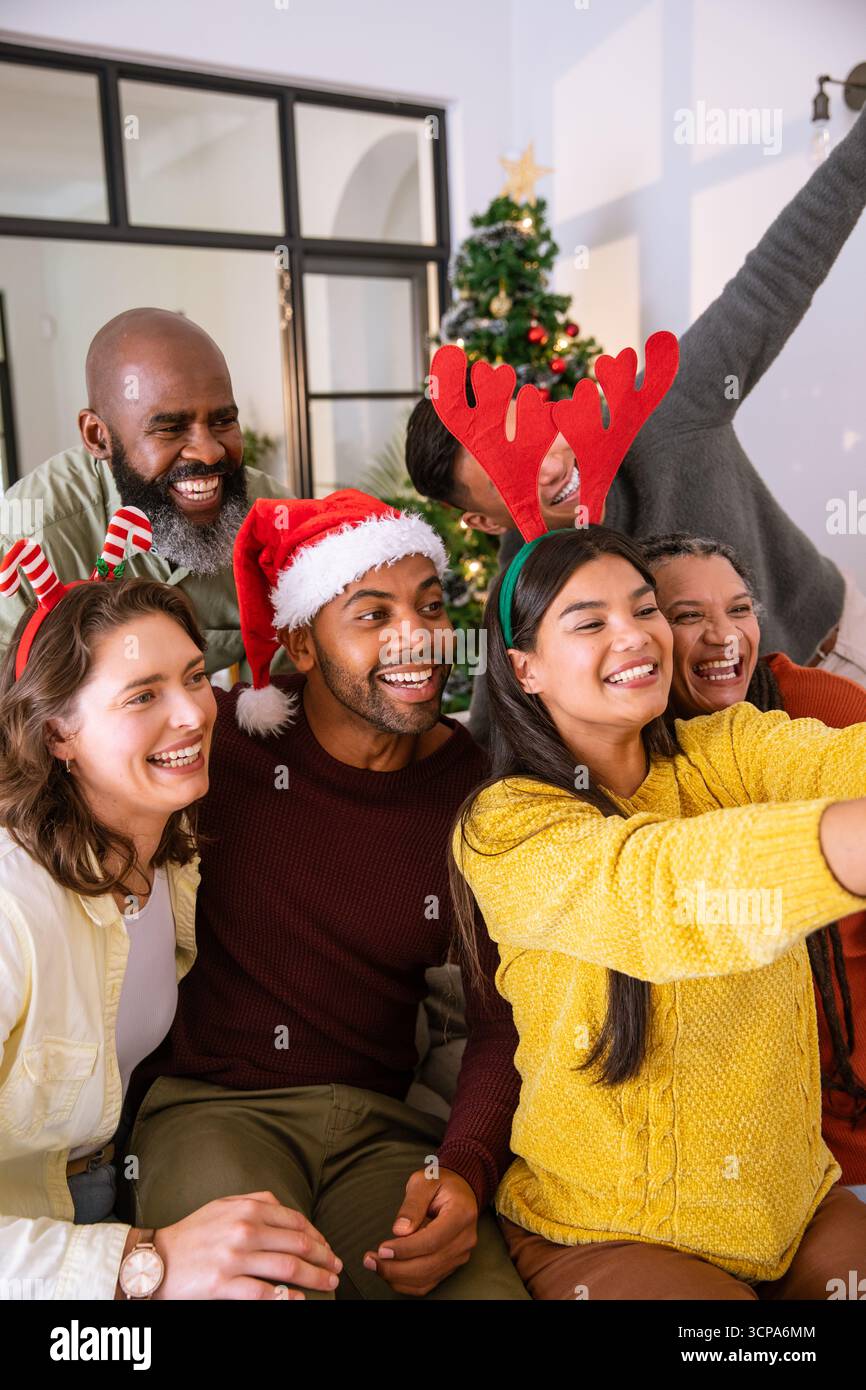 Amici diversi che si appoggiano sul divano di casa sotto l'albero di natale e scattano selfie con lo smartphone Foto Stock