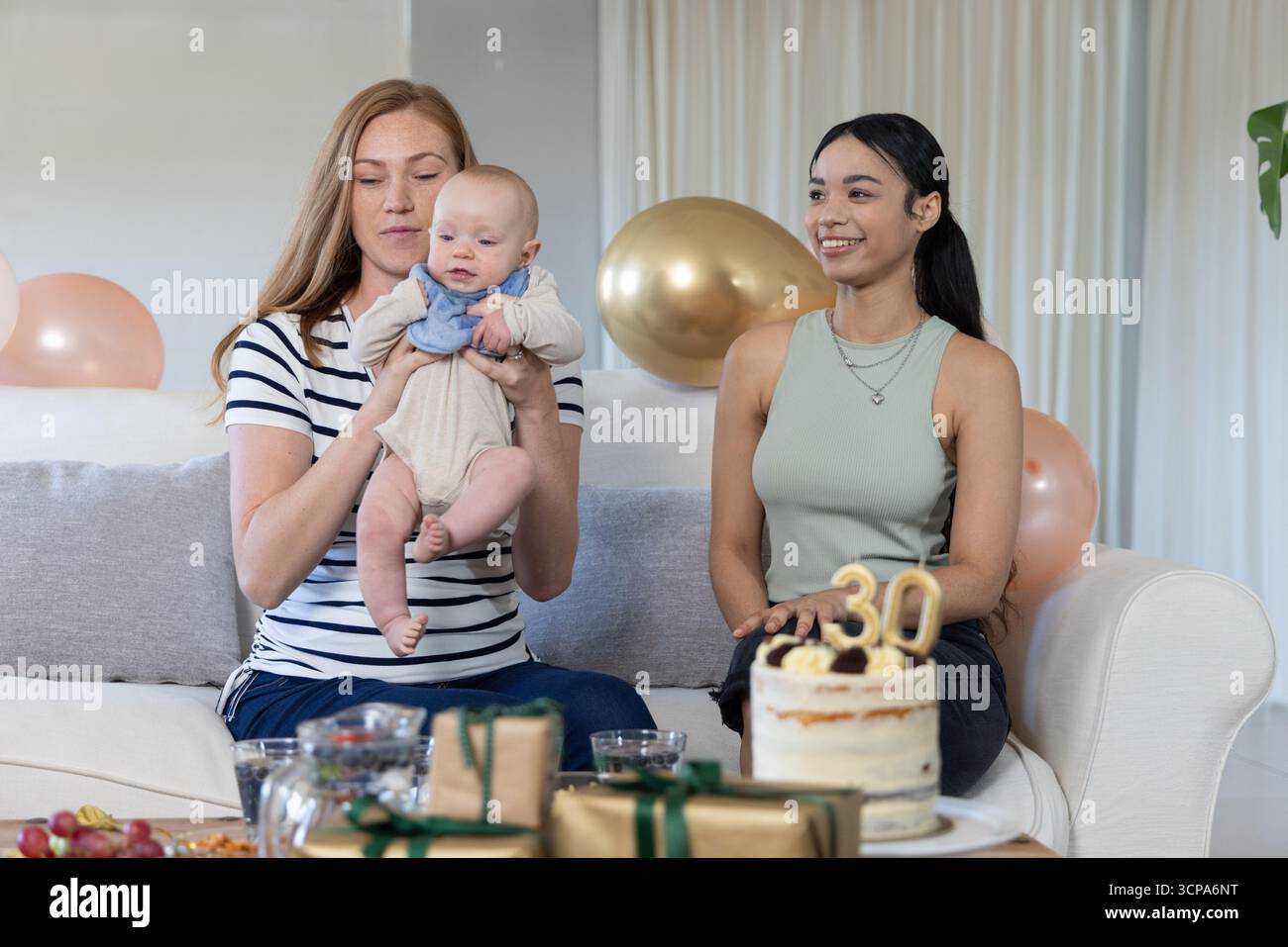 Diverse partner femminili e neonati che festeggiano il compleanno nel soggiorno sul divano con torta e regali Foto Stock