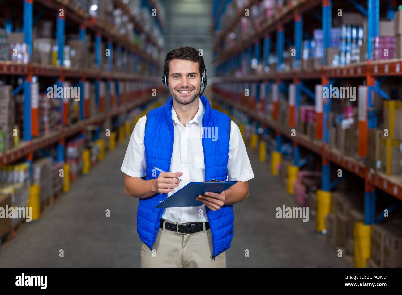 Lavoratore di magazzino in piedi nella corsia fiancheggiato da scaffali con scatole impilate, che reggono appunti Foto Stock