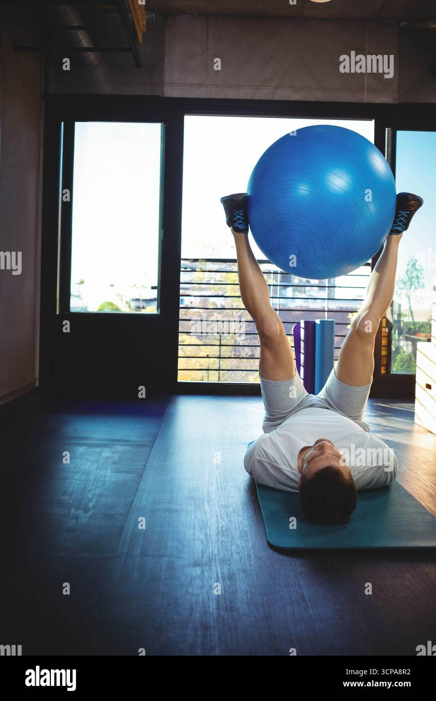 Uomo che indossa abiti da ginnastica che solleva palla blu di stabilità con gambe su tappetino per esercizi nella sala fitness Foto Stock