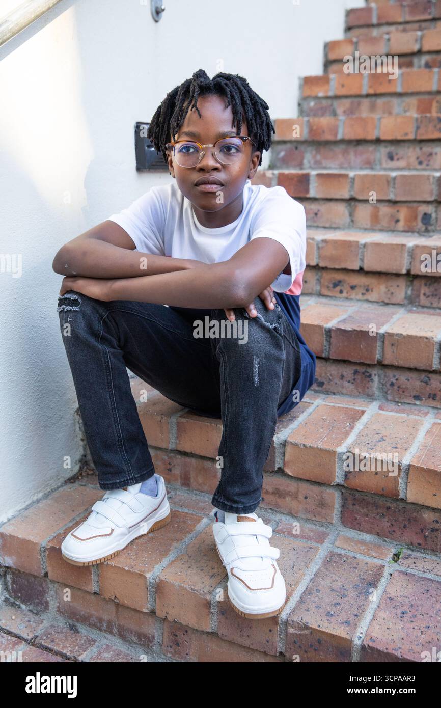 Adolescente afroamericano seduto su gradini di mattoni con occhiali e scarpe da ginnastica Foto Stock