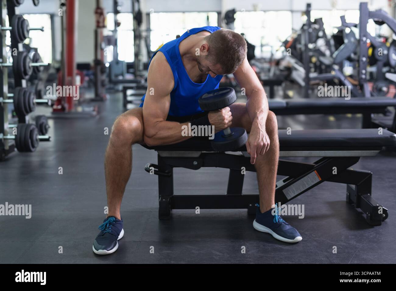 Uomo seduto sulla panchina di peso che esegue ricci in palestra indossando abbigliamento sportivo con manubrio Foto Stock