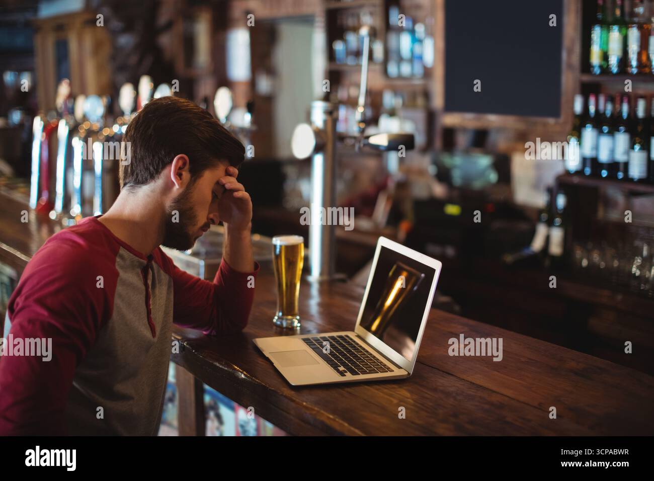Uomo barbuto che riposa a portata di mano mentre guarda il computer portatile accanto al bicchiere di birra al pub, copia dello spazio Foto Stock
