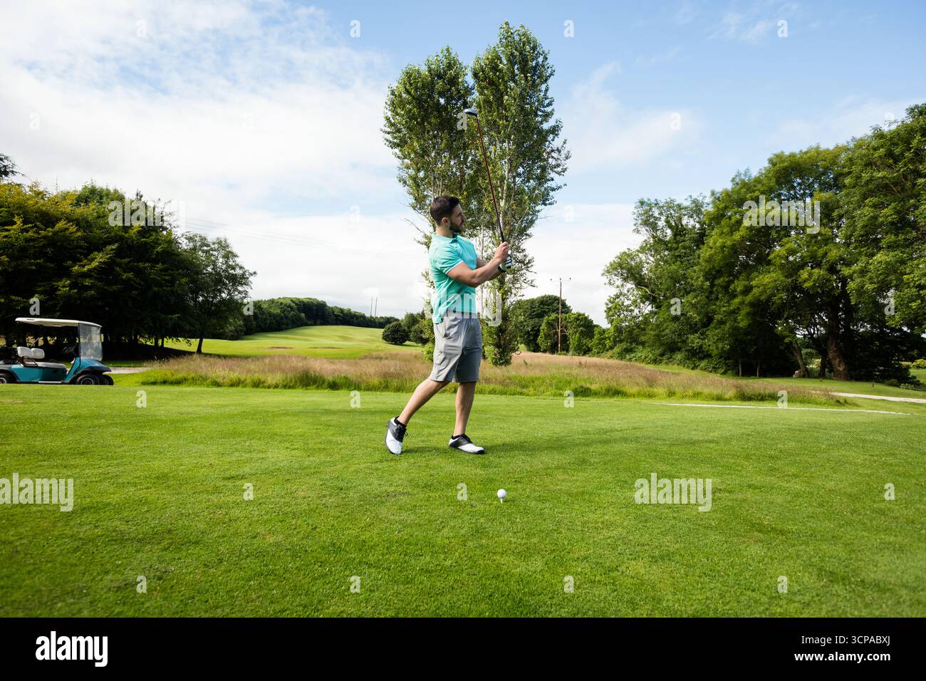 Uomo che oscilla sul tee box del campo da golf che si affaccia sul fairway con palla e cart Foto Stock