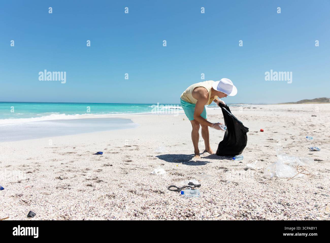 Volontario maschile che indossa un cappello da sole che raccoglie la bottiglia di plastica sulla spiaggia nel sacchetto della spazzatura, copia dello spazio Foto Stock