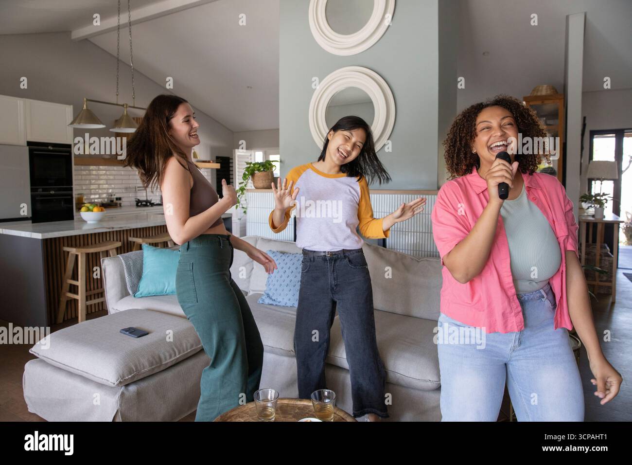 Diverse amiche che cantano, ballano nella cucina del soggiorno tenendo il microfono wireless vicino al divano Foto Stock