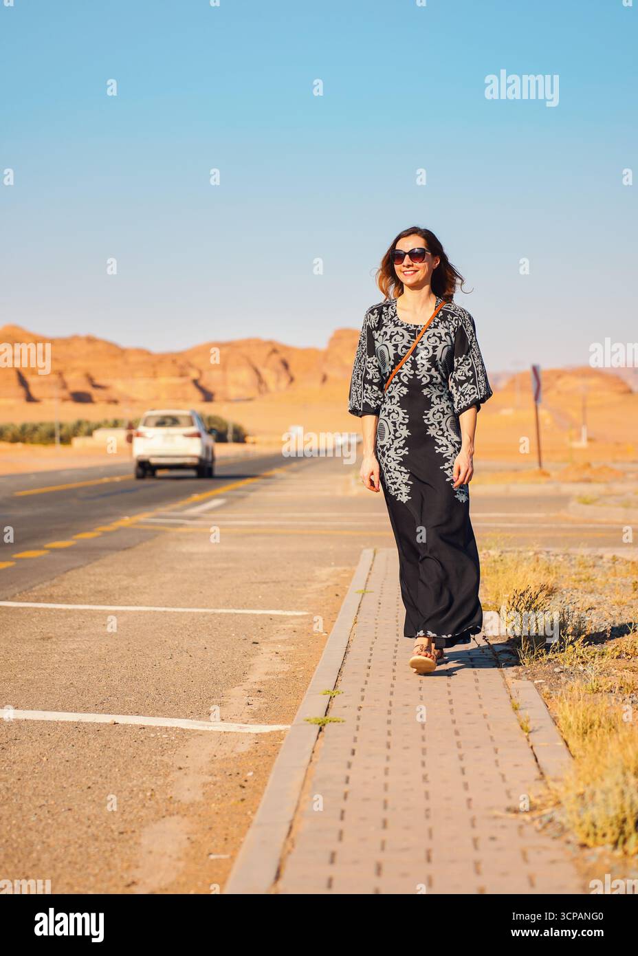 Giovane donna con lungo abito nero che cammina sul marciapiede vicino alla strada - paesaggio roccioso del deserto, tipico di Alula, sullo sfondo Foto Stock