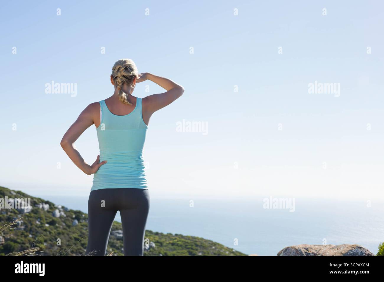 Donna in piedi su un punto panoramico roccioso con arbusti che ombreggiano gli occhi di fronte all'oceano in abbigliamento atletico, copia spazio Foto Stock