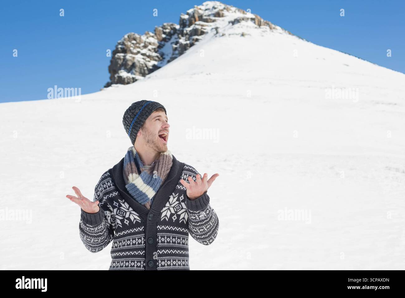 Uomo adulto che solleva le braccia su una pista innevata con maglione invernale e berretto in maglia di fronte alla vetta rocciosa Foto Stock