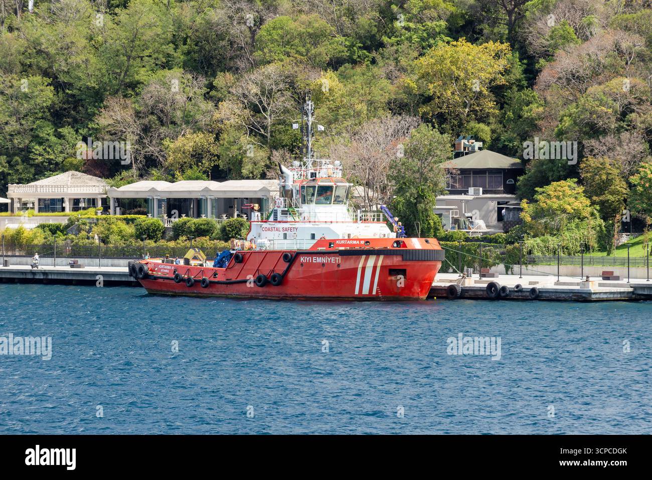 Istanbul, Turchia - 24 agosto 2024: Un rimorchiatore rosso brillante Kurtarma 10 è ormeggiato in un porto tranquillo accanto a un lungomare. Alberi lussureggianti e piccoli bu Foto Stock