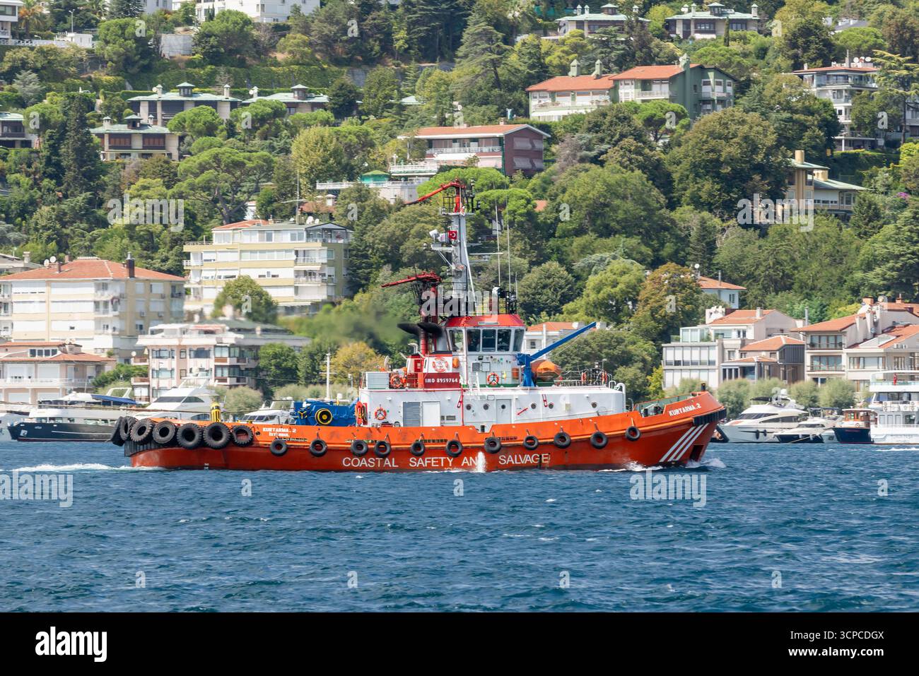 Istanbul, Turchia - 24 agosto 2024: Una nave di salvataggio arancione brillante etichettata Coastal Safety and Salvage si muove attraverso le acque blu calme, con una collina che conduce a. Foto Stock