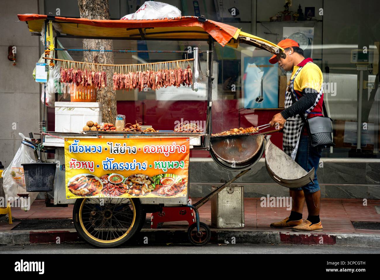 Bangkok, Thailandia, 5 settembre 2025: Un venditore cucina carni spiedate su un carrello di cibo di strada. E' decorata con immagini colorate di piatti. Foto Stock
