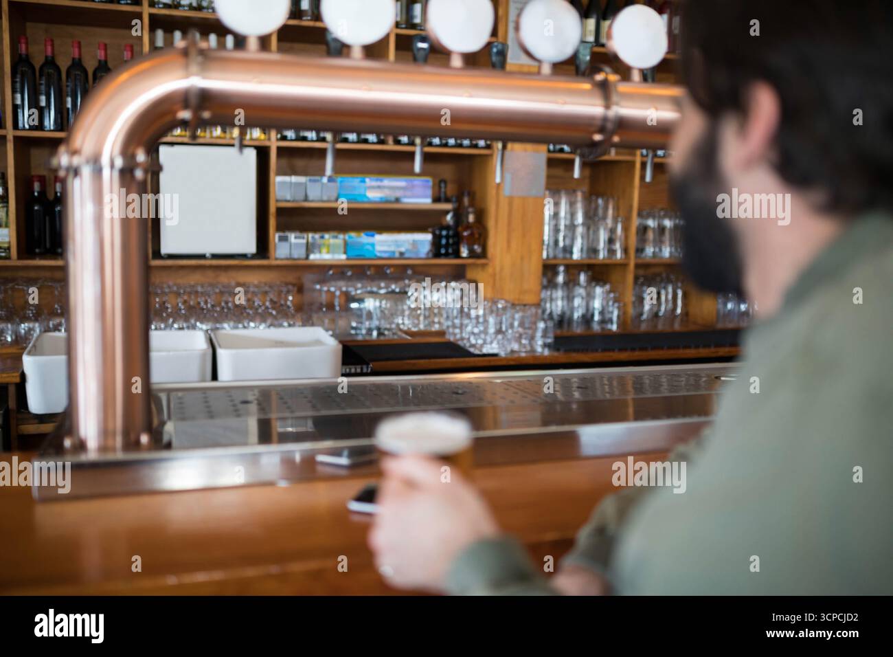 Uomo seduto al bar con una camicia verde oliva che tiene un bicchiere di birra vicino ai rubinetti di rame, spazio per le copie Foto Stock