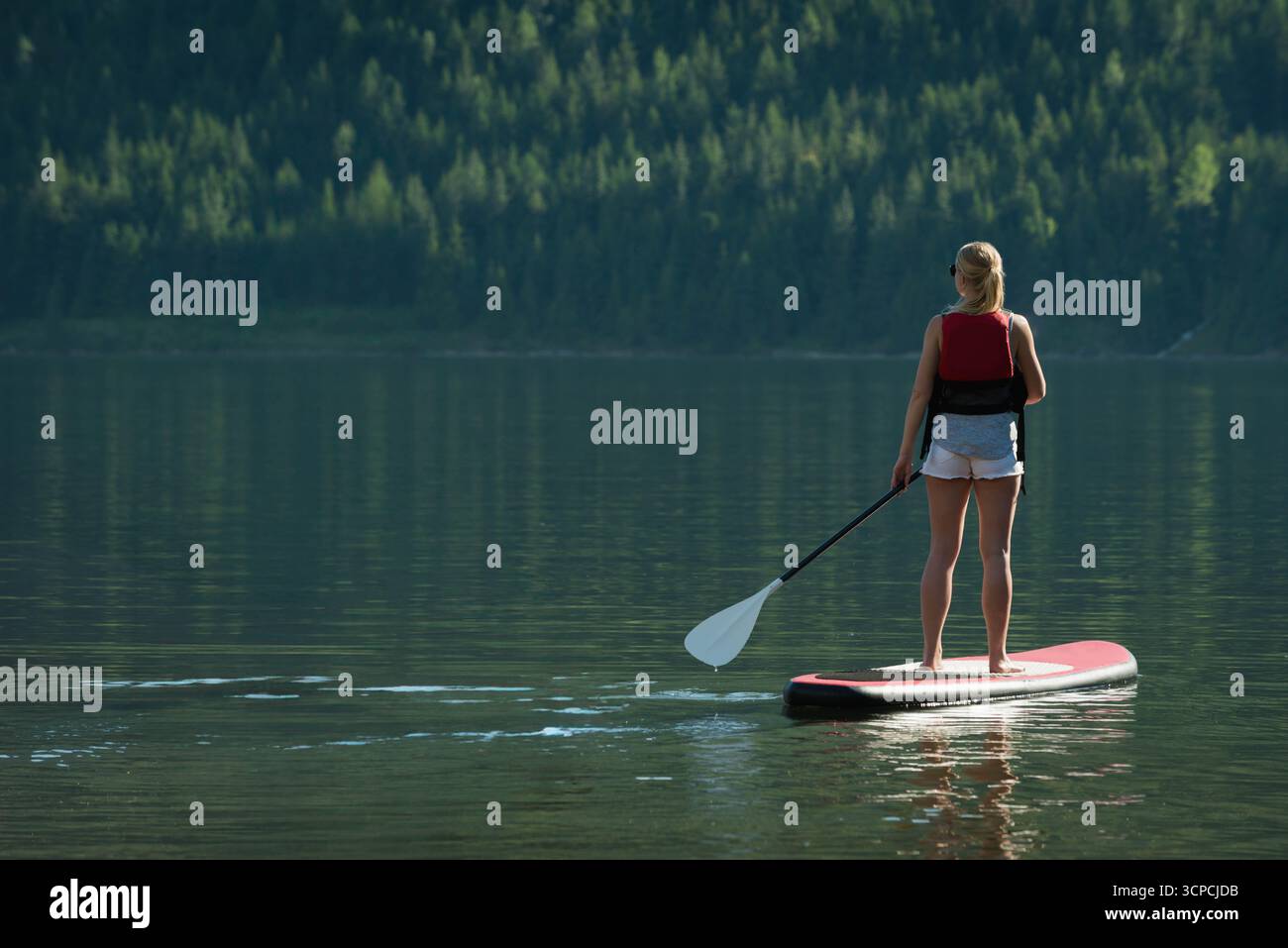 Paddleboarder donna che canta su paddleboard rosso in lago con gilet rosso, pantaloncini, spazio copia Foto Stock