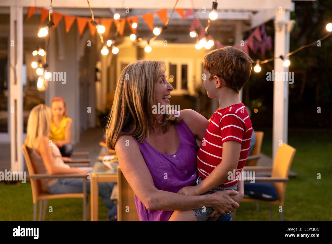 Riunione di famiglia multigenerazionale a tavola sotto pergola con luci a corda e accatastamento Foto Stock
