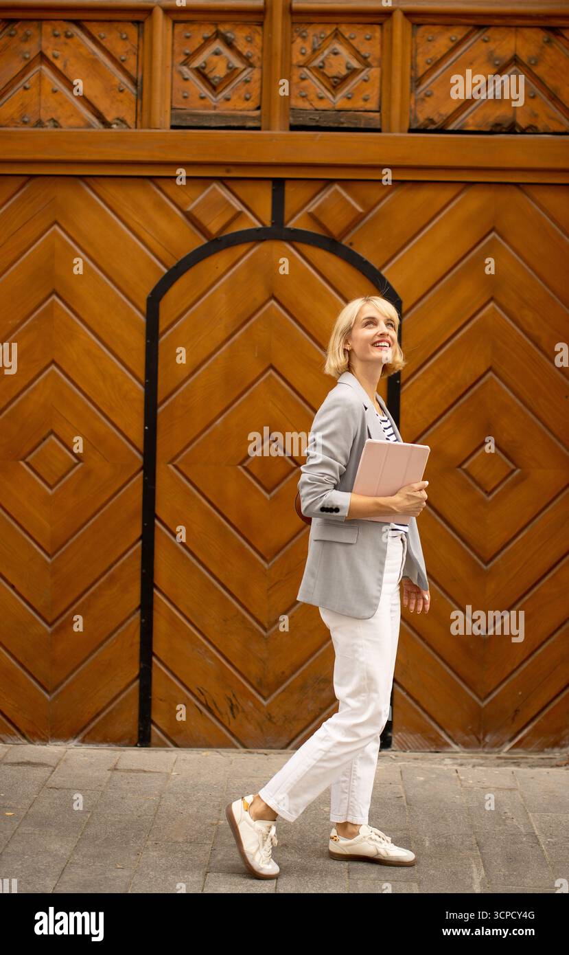 Una donna cammina con grazia tenendo in mano un notebook, godendosi una giornata di sole in città. Indossa un outfit elegante con un blazer e pantaloni bianchi, Foto Stock