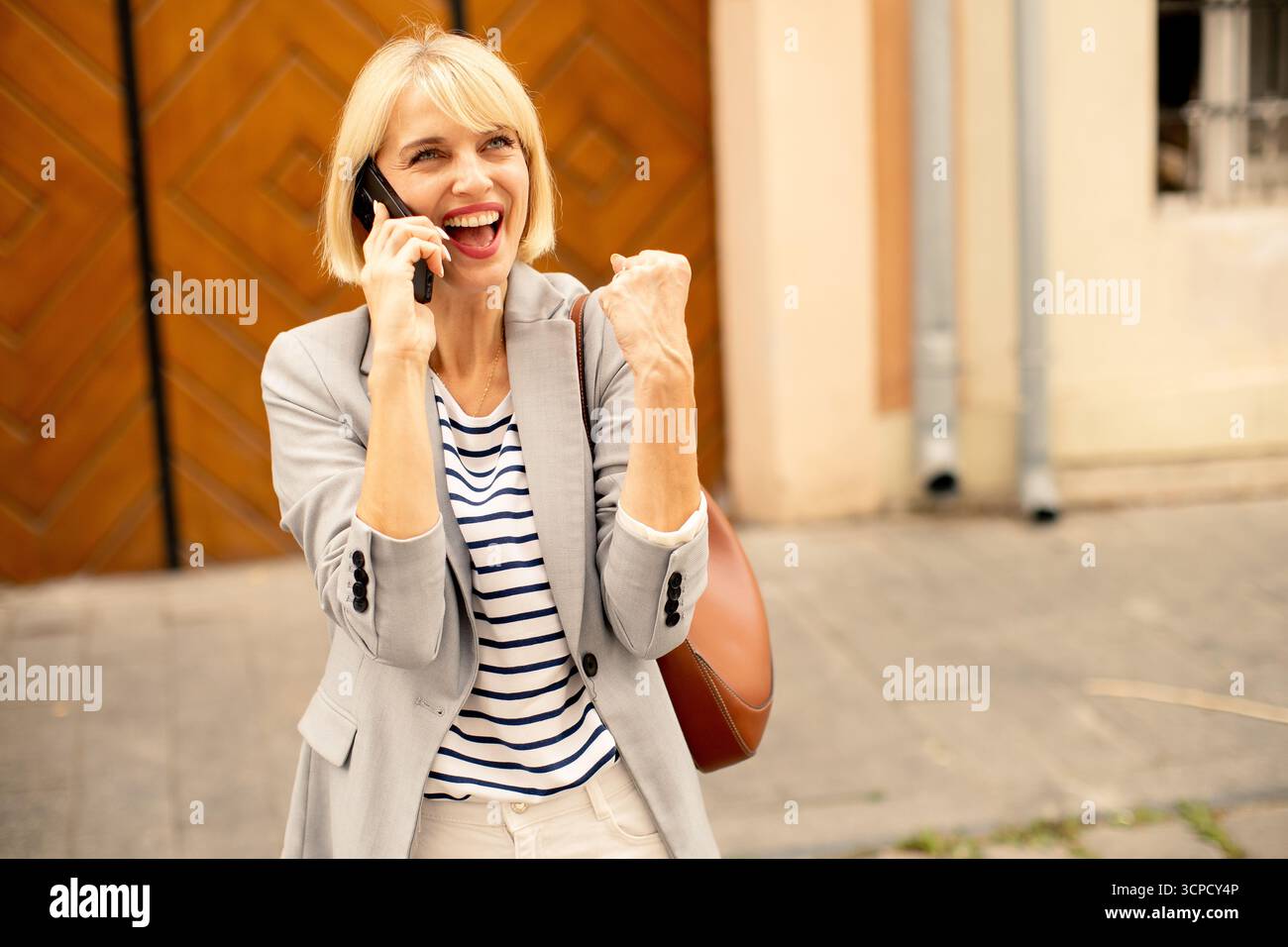 Una donna con i capelli biondi corti sorride in grande, alzando il pugno per festeggiare. Indossa un blazer grigio e parla al telefono fuori da un cha Foto Stock