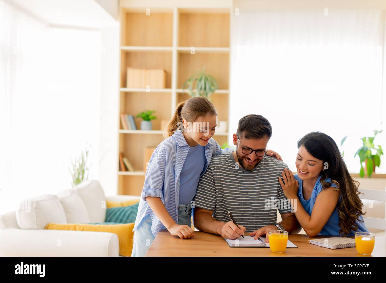 Una famiglia gioiosa si impegna in attività creative insieme mentre si gusta un succo di frutta fresco. Le Risate riempiono lo spazio illuminato dal sole mentre condividono i momenti. Foto Stock