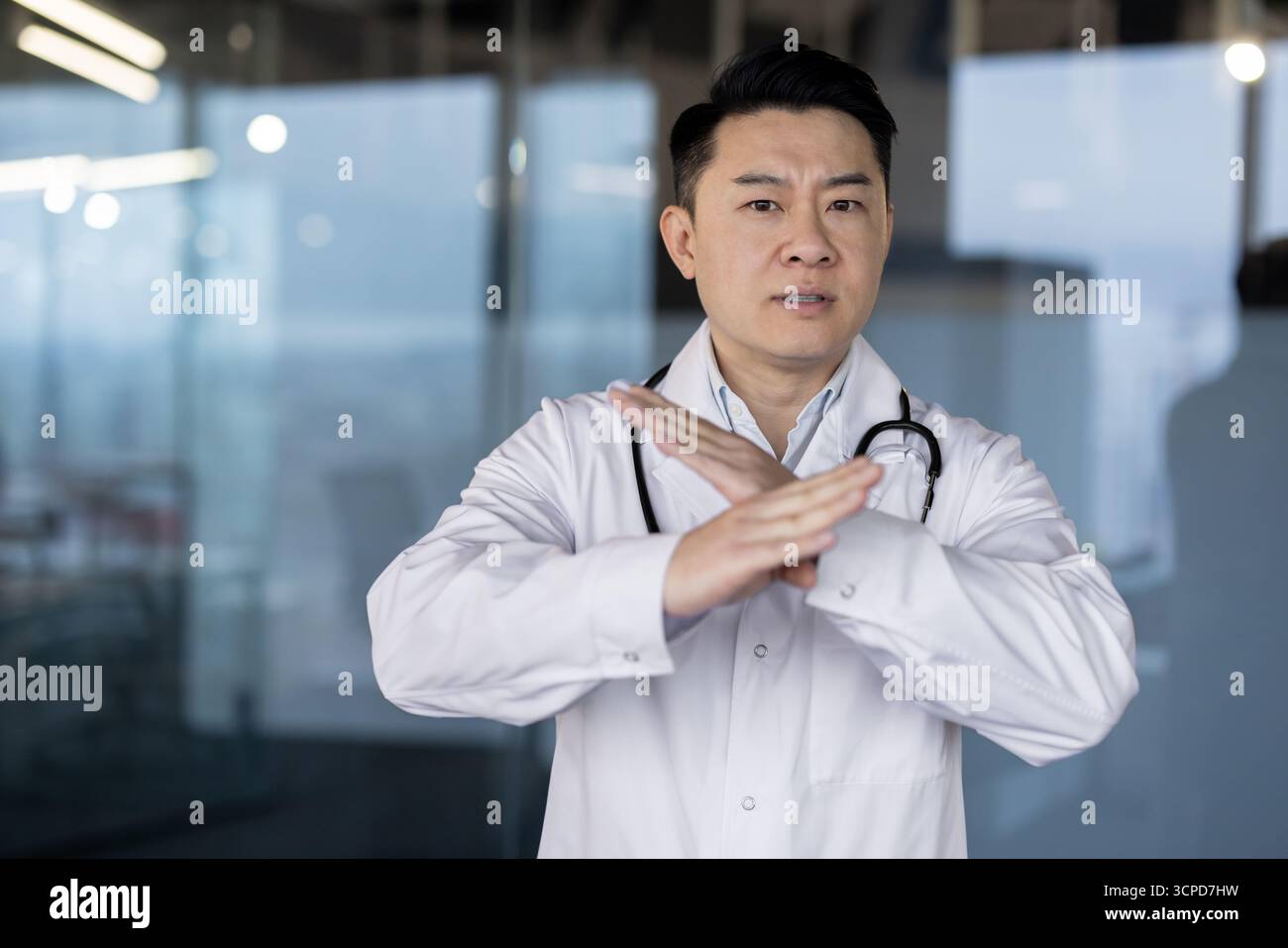 Ritratto di un medico asiatico giovane serio e preoccupato in un cappotto bianco in piedi nell'atrio della clinica e che mostra un no stop con le mani alla macchina fotografica. Foto Stock