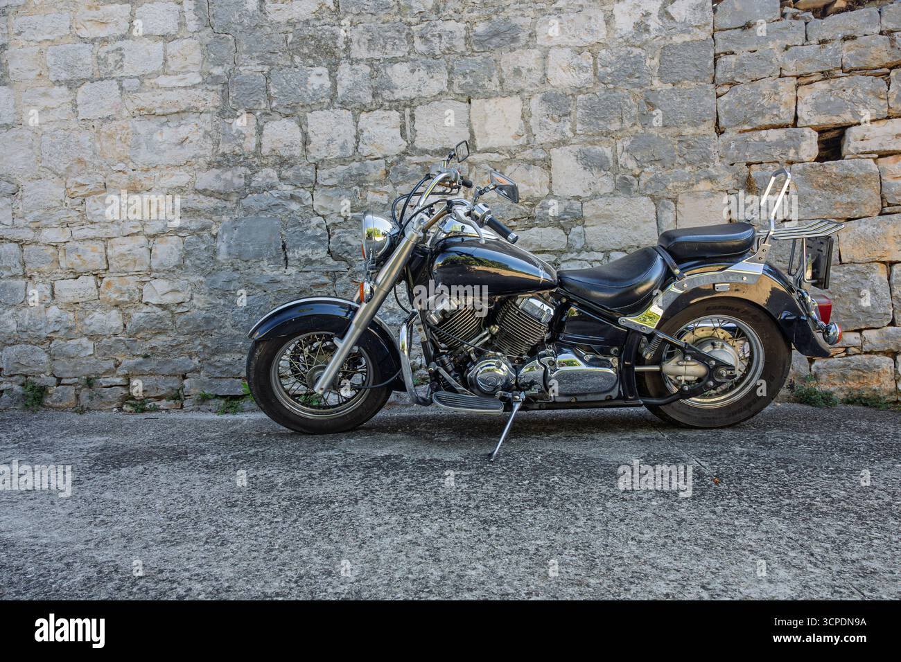 Una classica motocicletta cruiser nera parcheggiata accanto a una rustica parete in pietra, con accenti cromati e stile vintage. Semplice, robusto e senza tempo: Perfetto per te Foto Stock