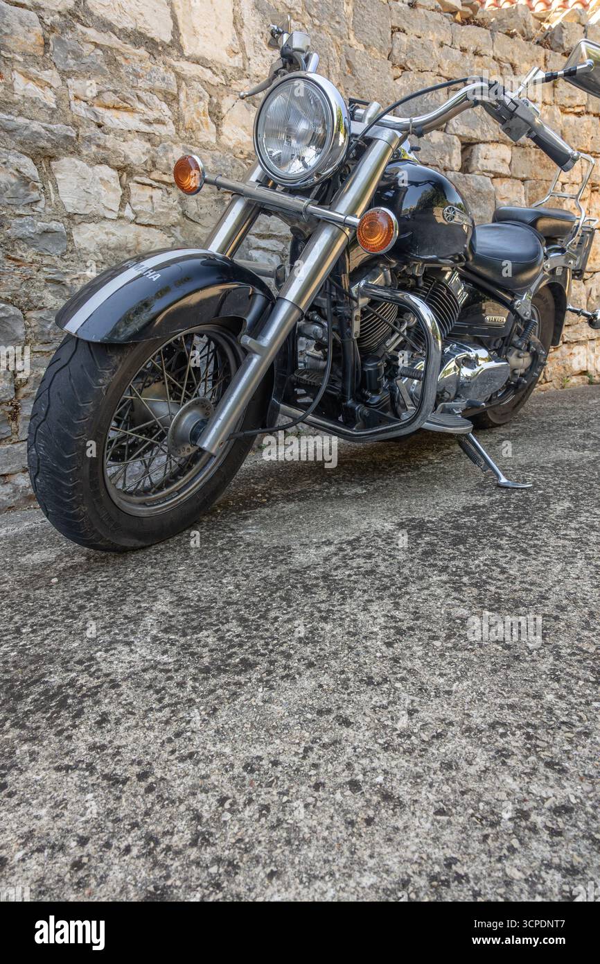 Una classica motocicletta cruiser nera parcheggiata accanto a una rustica parete in pietra, con accenti cromati e stile vintage. Semplice, robusto e senza tempo: Perfetto per te Foto Stock